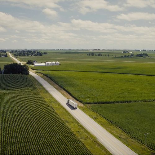 An image of farm lands as far as the eye can see with a wide blue sky and a semi-truck on the road.