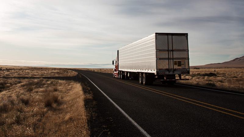 A semi-truck driving down a road into the sunset.